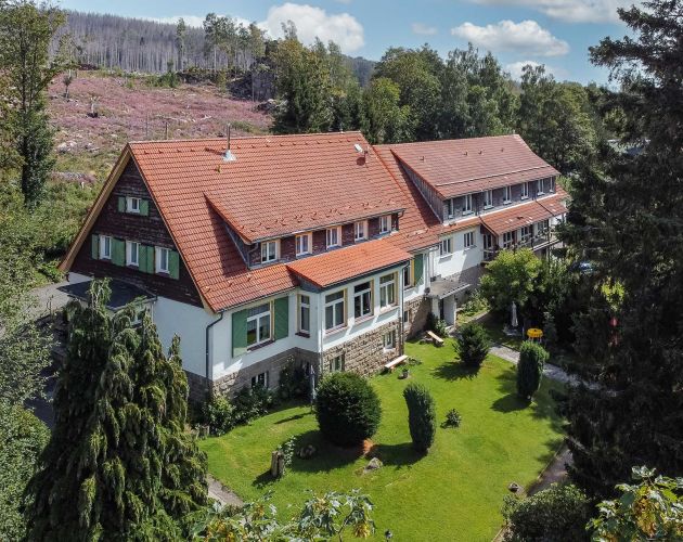Landhaus mit rotem Dach und weißer Fassade, umgeben von Gärten und Wald.