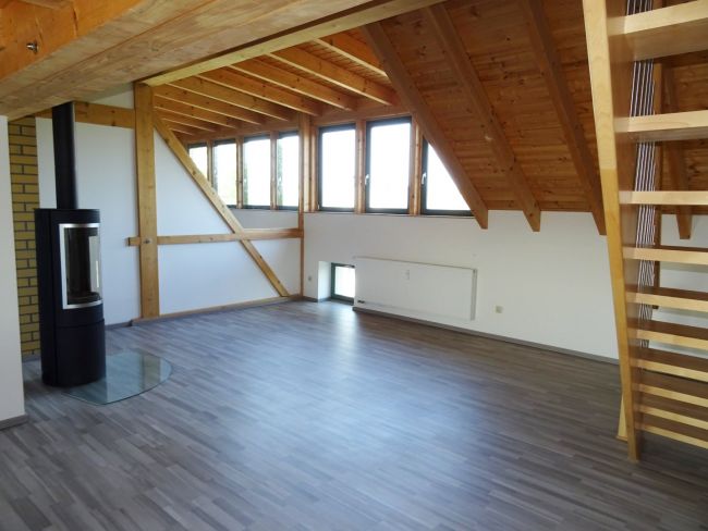 Helles Dachgeschoss mit Holzbalken, Kaminofen und moderner Treppe – offener Wohnbereich mit großen Fenstern.