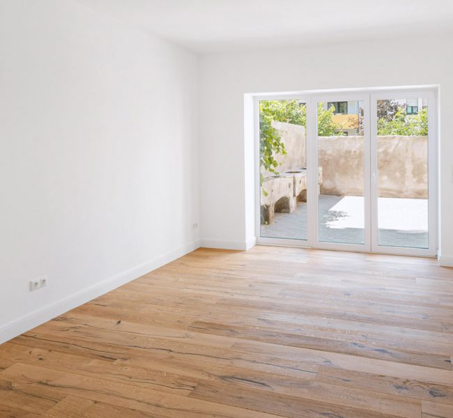 Helles Zimmer mit Holzfußboden und großer Terrassentür mit Blick auf einen Innenhof.