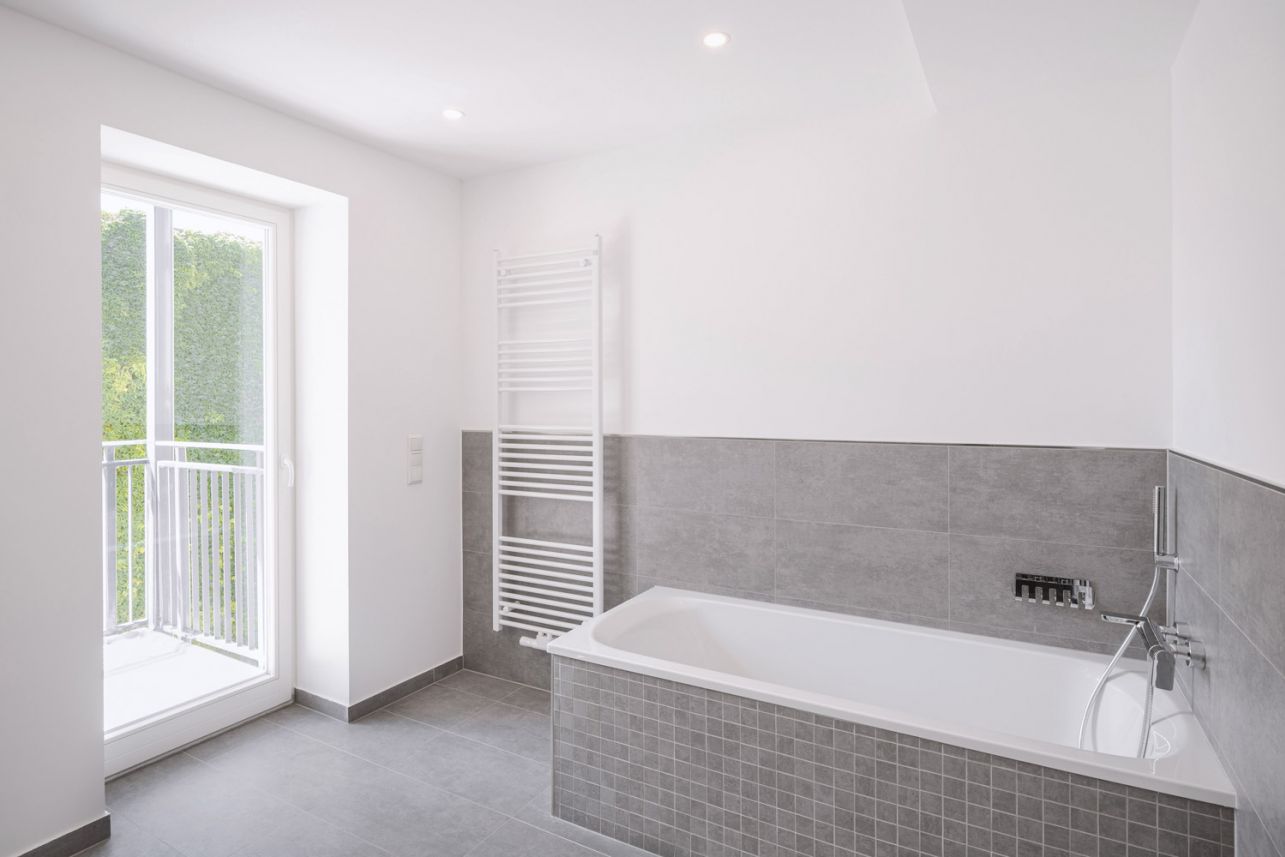 Modernes Badezimmer mit grauen Fliesen, Badewanne und bodentiefem Fenster zum Balkon.