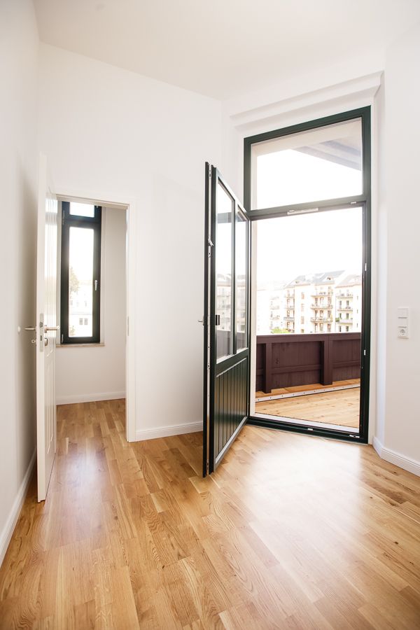 Helles Zimmer mit Holzboden, hoher Decke und bodentiefem Fenster mit Zugang zu einem Balkon mit Blick auf Wohnhäuser.