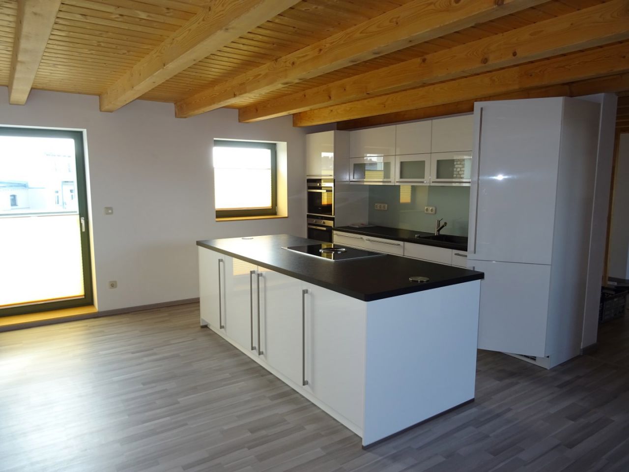 Moderne offene Küche mit Kochinsel, weißen Hochglanzfronten und Holzdecke in einem lichtdurchfluteten Wohnraum.