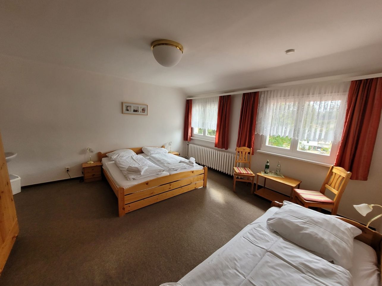 Gemütliches Gästezimmer mit Holzmöbeln, drei Betten, großen Fenstern und warmen Vorhängen – einladende Unterkunft für Erholung.