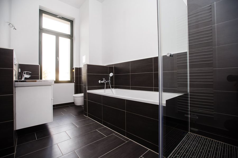 Modernes Badezimmer mit schwarzer Fliesenoptik, Badewanne, Glasdusche und großem Fenster für viel Tageslicht.