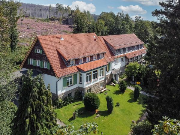 Landhaus mit rotem Dach und weißer Fassade, umgeben von Gärten und Wald.