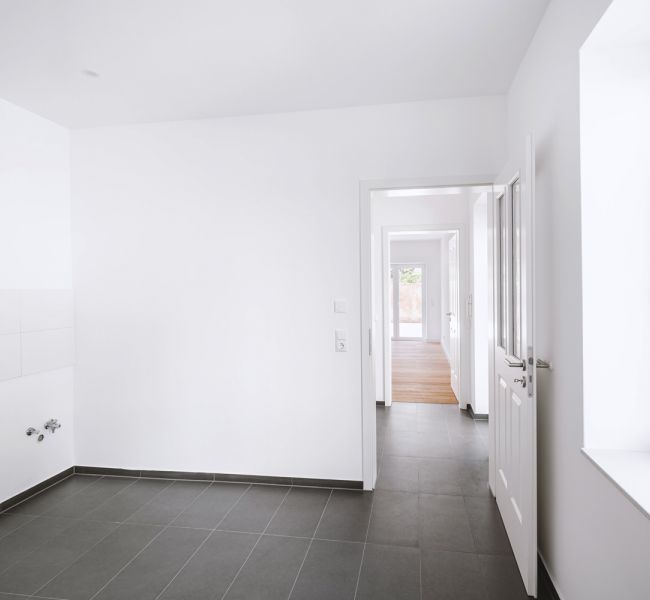 Helle, leere Küche mit weißen Wänden, grauen Fliesen und großem Fenster.