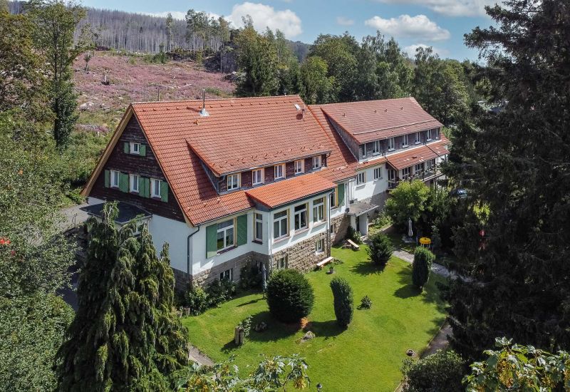 Landhaus mit rotem Dach und weißer Fassade, umgeben von Gärten und Wald.
