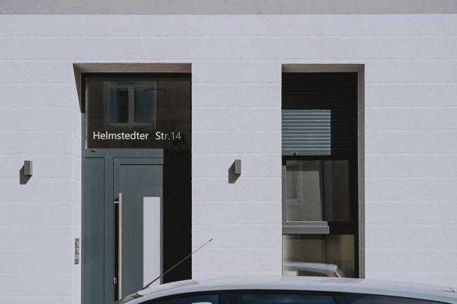 Moderner Hauseingang mit dunkler Tür und Glasbeschriftung „Helmstedter Str. 14“ an einer weißen Fassade.