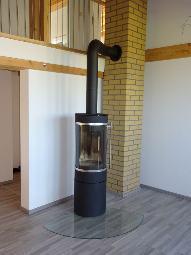 Moderner Kaminofen mit Glasplatte vor gemauerter Wand in einem Wohnraum mit Holz-Elementen und offener Galerie.