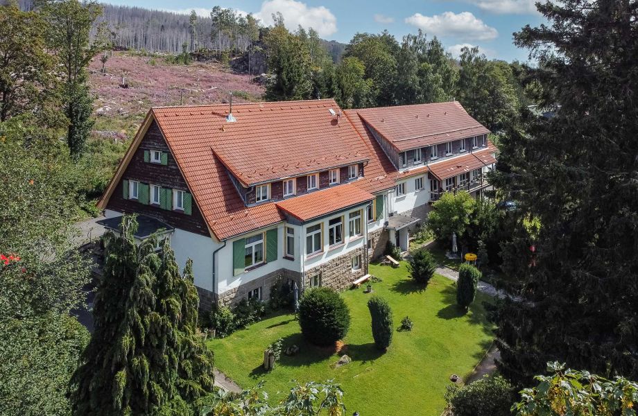 Landhaus mit rotem Dach und weißer Fassade, umgeben von Gärten und Wald.